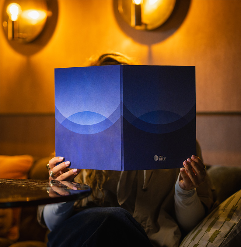 Persoon in Bar Blue Nijmegen die een blauw menu met het logo van bar BLUE voor het gezicht houdt.